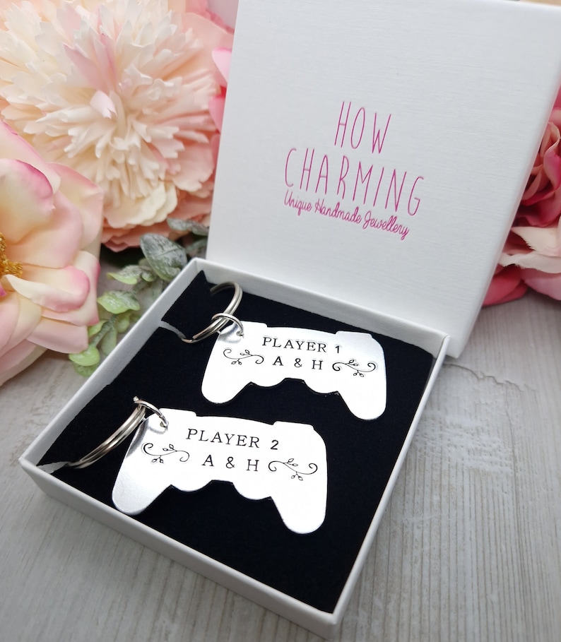 Op de afbeelding: Twee zilveren sleutelhangers in de vorm van videogamecontrollers met de tekst "PLAYER 1 A&H" en "PLAYER 2 A&H" erop. De sleutelhangers zitten in een witte doos met een zwarte binnenkant.