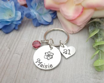 May include: Silver keychain with a round charm engraved with the name "Maisie" and a small flower design. A smaller heart-shaped charm is attached to the ring, engraved with the number "21". A small pink crystal is attached to the ring.