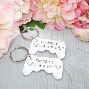 Op de afbeelding: Twee zilveren sleutelhangers in de vorm van videogamecontrollers. De bovenste sleutelhanger zegt "Player 1 A & H" en de onderste sleutelhanger zegt "Player 2 A & H".