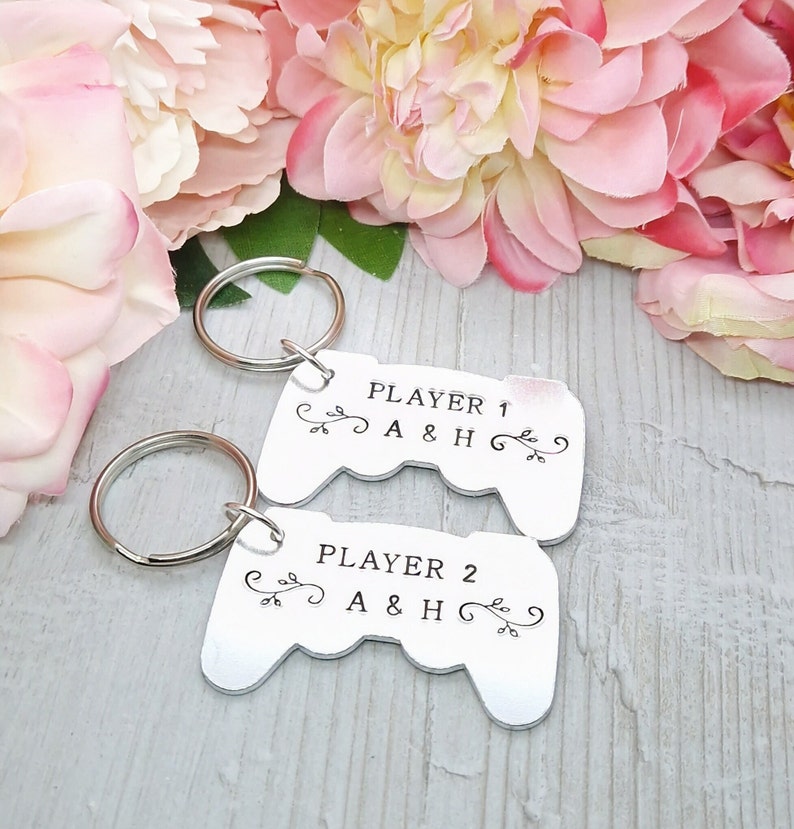 Op de afbeelding: Twee zilveren sleutelhangers in de vorm van videogamecontrollers. De bovenste sleutelhanger zegt "Player 1 A & H" en de onderste sleutelhanger zegt "Player 2 A & H".