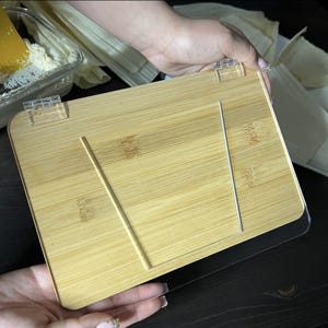 May include: A bamboo cookbook stand with a clear acrylic base and hinges. The stand has a light brown color and features a groove to hold a book open. The stand is designed to hold a cookbook or tablet.