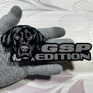 May include: A black and silver GSP Edition sticker featuring a detailed illustration of a dog's head. The text "GSP EDITION" is in silver block letters. The sticker is held by a gray glove.