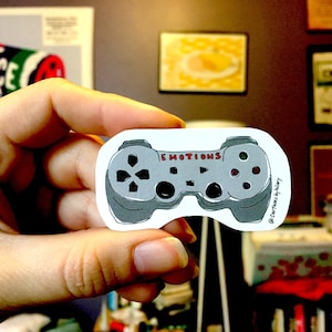 Op de afbeelding: Een grijze en witte sticker van een videogamecontroller met het woord "EMOTIONS" erop gedrukt. De sticker wordt in een hand gehouden.