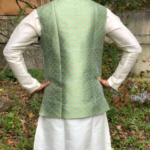 Peut inclure: Un gilet vert menthe avec un motif dor&eacute;. Le gilet est port&eacute; sur une chemise blanche et une longue jupe blanche.