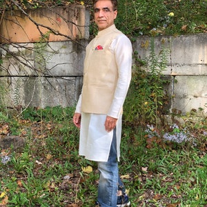 May include: A man wearing a beige waistcoat over a white shirt and blue jeans. He is standing in front of a stone wall with greenery behind him.