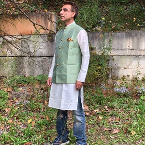 Peut inclure: Un homme portant un gilet &agrave; motifs vert et or sur une chemise blanche et un jean bleu. Il se tient dans une zone herbeuse avec un mur de pierre derri&egrave;re lui.