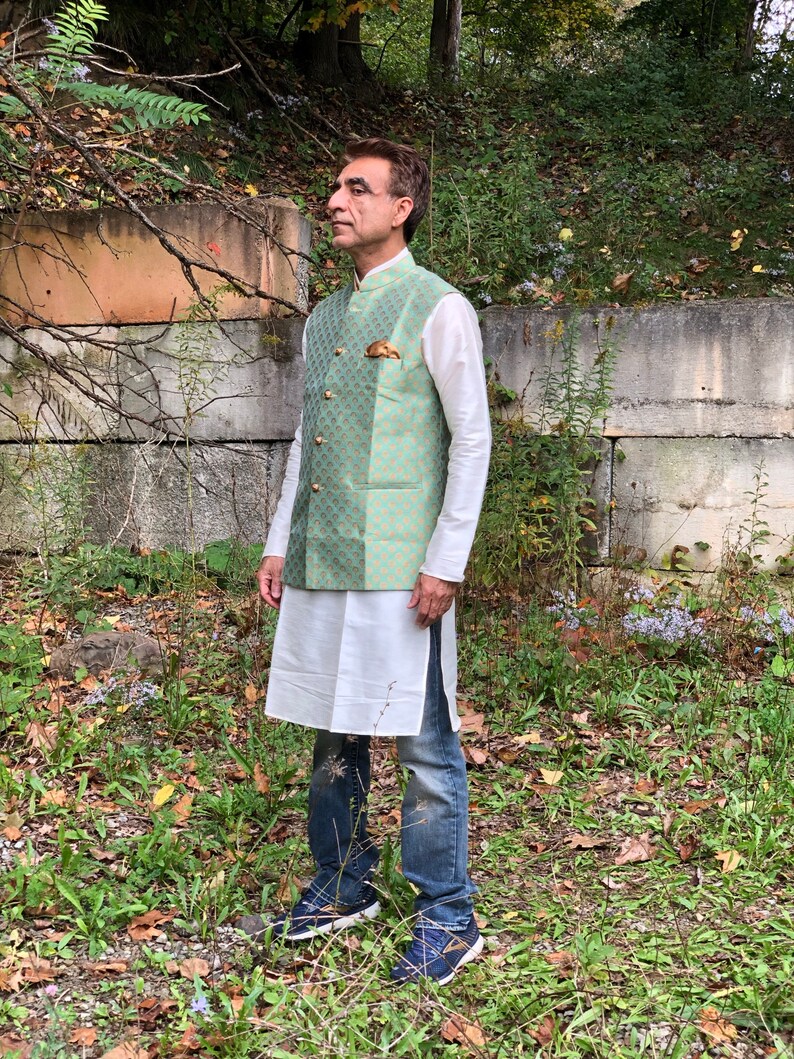 Peut inclure: Un homme portant un gilet &agrave; motifs vert et or sur une chemise blanche et un jean bleu. Il se tient dans une zone herbeuse avec un mur de pierre derri&egrave;re lui.