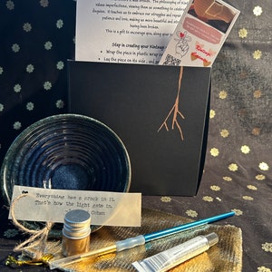 May include: A Kintsugi art kit with a blue ceramic bowl, gold paint, a brush, and a small bottle of gold leaf. The kit comes with instructions on how to repair broken pottery with gold. The text on the card reads "Everything has a crack in it. That's how the light gets in. - Leonard Cohen"