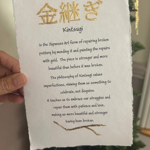 May include: A handmade paper with the Japanese word "Kintsugi" in gold lettering. The text describes Kintsugi as the art of repairing broken pottery with gold, emphasizing embracing imperfections. The paper is held in front of a Christmas tree.