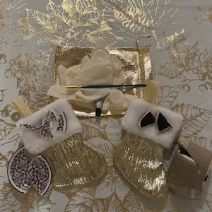 May include: Two gold Christmas stockings with white fur trim and a purple and white ceramic ornament on each stocking. The stockings are on a white tablecloth with a gold leaf pattern.