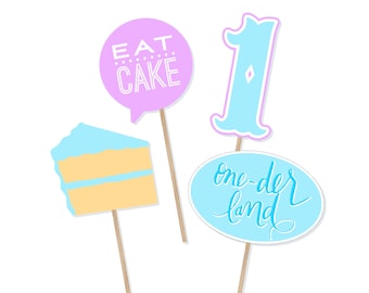Könnte beinhalten: Vier blaue und rosa Fotobooth-Requisiten mit Stöcken. Die Requisiten beinhalten ein Stück Kuchen, die Nummer eins, eine Sprechblase mit der Aufschrift "Eat Cake" und eine Sprechblase mit der Aufschrift "one-der land".
