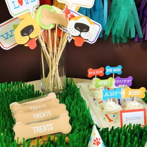 Puede incluir: Una mesa de fiesta con tem&aacute;tica de cachorros con papel verde parecido a la hierba, cupcakes con adornos con tem&aacute;tica de perros y una variedad de accesorios para cabina de fotos, incluyendo un cartel que dice "Puppy Love" y una nariz y lengua de perro.