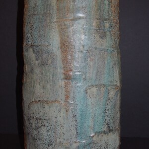 May include: A tall, cylindrical ceramic vase with a textured surface. The vase features a blend of blue, green, and brown hues, with a rough, weathered appearance. The design suggests a handmade, artisanal quality, suitable for home decor.