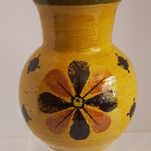May include: A yellow ceramic vase with a round body and a flared neck. The vase features a large floral design in brown and orange, with smaller black accents. The rim is trimmed with green.