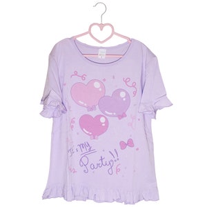 May include: A lavender t-shirt with a ruffled hem and short sleeves. The shirt features a graphic of three pink and purple heart-shaped balloons with bows and the text "It's my Party!" in pink.