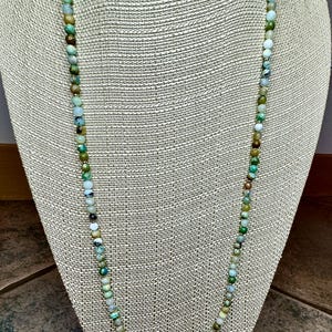 May include: A long necklace made of green, blue, and white gemstone beads. The necklace is displayed on a fabric-covered mannequin.