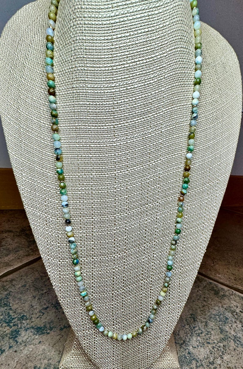 May include: A long necklace made of green, blue, and white gemstone beads. The necklace is displayed on a fabric-covered mannequin.