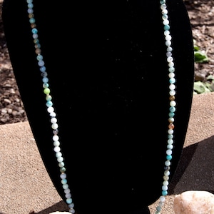 May include: A long necklace made of various shades of green, blue, and white beads. The necklace is displayed on a black velvet mannequin.