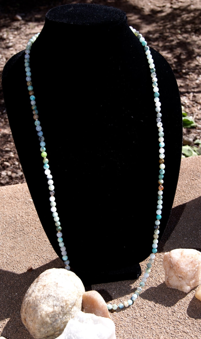 May include: A long necklace made of various shades of green, blue, and white beads. The necklace is displayed on a black velvet mannequin.