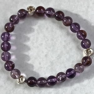 Könnte beinhalten: Ein Armband aus violetten Amethyst-Edelsteinen mit silbernen Akzenten. Das Armband besteht aus runden Perlen und hat einen Verschluss.