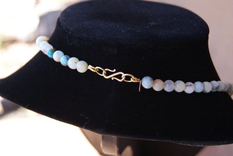 May include: A close-up of a light blue gemstone necklace with a gold clasp. The necklace is displayed on a black velvet stand.