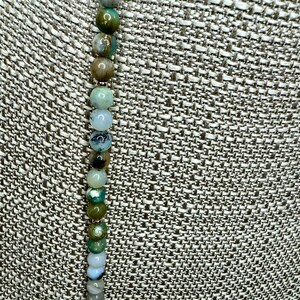 May include: A strand of green, brown, and white agate beads on a fabric background.