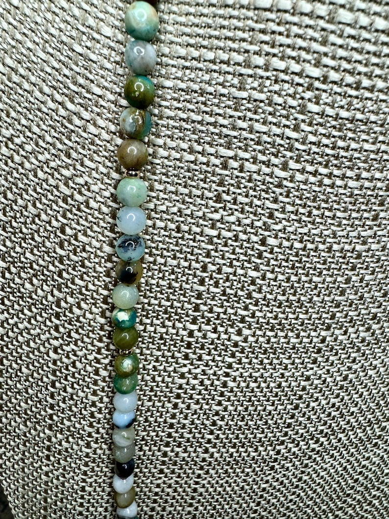May include: A strand of green, brown, and white agate beads on a fabric background.