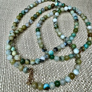May include: A long necklace made of green, brown, and white gemstone beads. The necklace has a gold clasp.