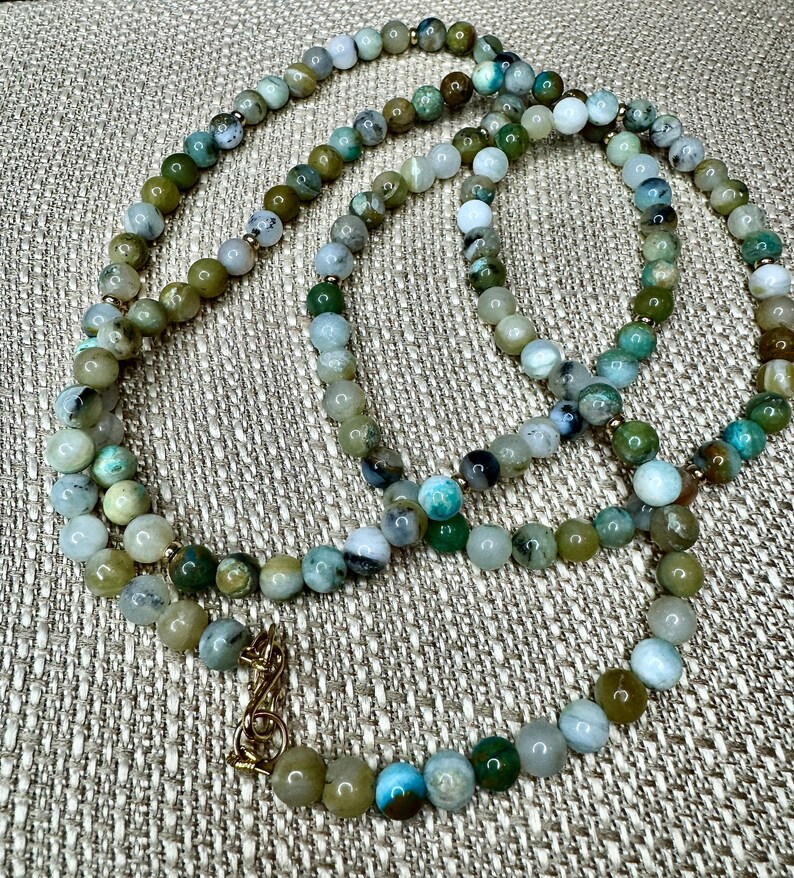 May include: A long necklace made of green, brown, and white gemstone beads. The necklace has a gold clasp.