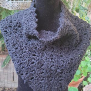 May include: A dark gray crocheted cowl scarf displayed on a black mannequin. The scarf has a textured, open weave design and a scalloped edge. The cowl is designed to be worn around the neck and shoulders.