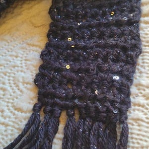 May include: Dark navy blue crocheted scarf with silver sequins. The scarf has a textured, chunky knit design and long fringe tassels at the ends. The scarf is handmade and appears soft and warm, suitable for cold weather.