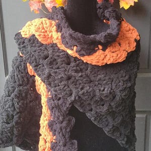 May include: A handmade, chunky knit shawl in black and orange, displayed on a mannequin. The shawl features a contrasting orange trim and a matching cowl neck. The background includes a fall wreath with sunflowers and autumn leaves.