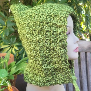 May include: A handmade green crochet hood with pointed ears. The hood is a mix of light and dark green yarn, creating a textured appearance. The hood has a drawstring and is displayed on a mannequin head.