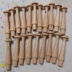 May include: A collection of nineteen light brown wooden pegs arranged in two rows. Each peg features a rounded top, a tapered cylindrical body, and a smaller cylindrical base designed for insertion. The wood grain is visible on each piece.