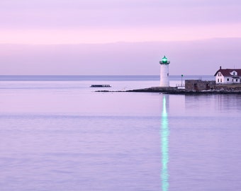 May include: A serene coastal scene featuring a white lighthouse with a green light atop, next to a white house with a red roof. The lighthouse's reflection shimmers in the calm, lavender-colored water. The sky is a soft lavender hue.