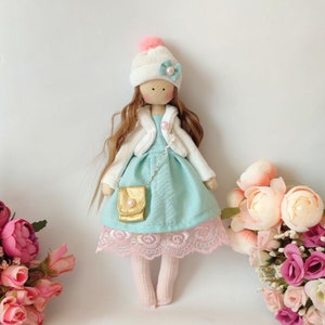 Puede incluir: Una mu&ntilde;eca hecha a mano con cabello largo casta&ntilde;o, que lleva un sombrero blanco con un pomp&oacute;n rosa, una chaqueta blanca, un vestido azul claro con un borde de encaje rosa y calcetines blancos. La mu&ntilde;eca sostiene un peque&ntilde;o bolso dorado y est&aacute; de pie sobre un fondo blanco.