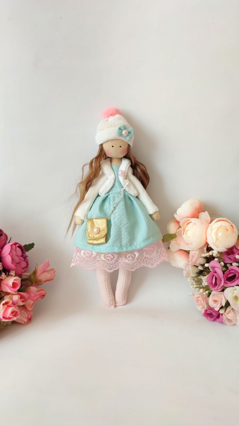 Puede incluir: Una mu&ntilde;eca hecha a mano con cabello largo casta&ntilde;o, que lleva un sombrero blanco con un pomp&oacute;n rosa, una chaqueta blanca, un vestido azul claro con un borde de encaje rosa y calcetines blancos. La mu&ntilde;eca sostiene un peque&ntilde;o bolso dorado y est&aacute; de pie sobre un fondo blanco.