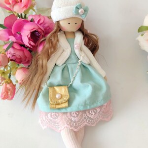 Puede incluir: Una mu&ntilde;eca hecha a mano con cabello largo y casta&ntilde;o, que lleva un sombrero blanco con un pomp&oacute;n rosa, una chaqueta blanca, un vestido azul claro con un bolso dorado y calcetines rosas.