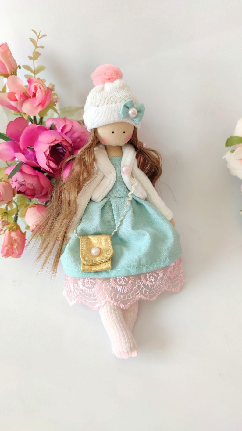 Puede incluir: Una mu&ntilde;eca hecha a mano con cabello largo y casta&ntilde;o, que lleva un sombrero blanco con un pomp&oacute;n rosa, una chaqueta blanca, un vestido azul claro con un bolso dorado y calcetines rosas.