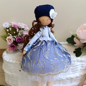 May include: A handmade doll with brown curly hair, wearing a blue and white floral dress with a blue hat and a white flower.