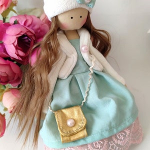 Puede incluir: Una mu&ntilde;eca hecha a mano con cabello largo y casta&ntilde;o, que lleva un gorro blanco de punto con un pomp&oacute;n rosa, una chaqueta blanca, un vestido azul claro con un bolso dorado y calcetines blancos.