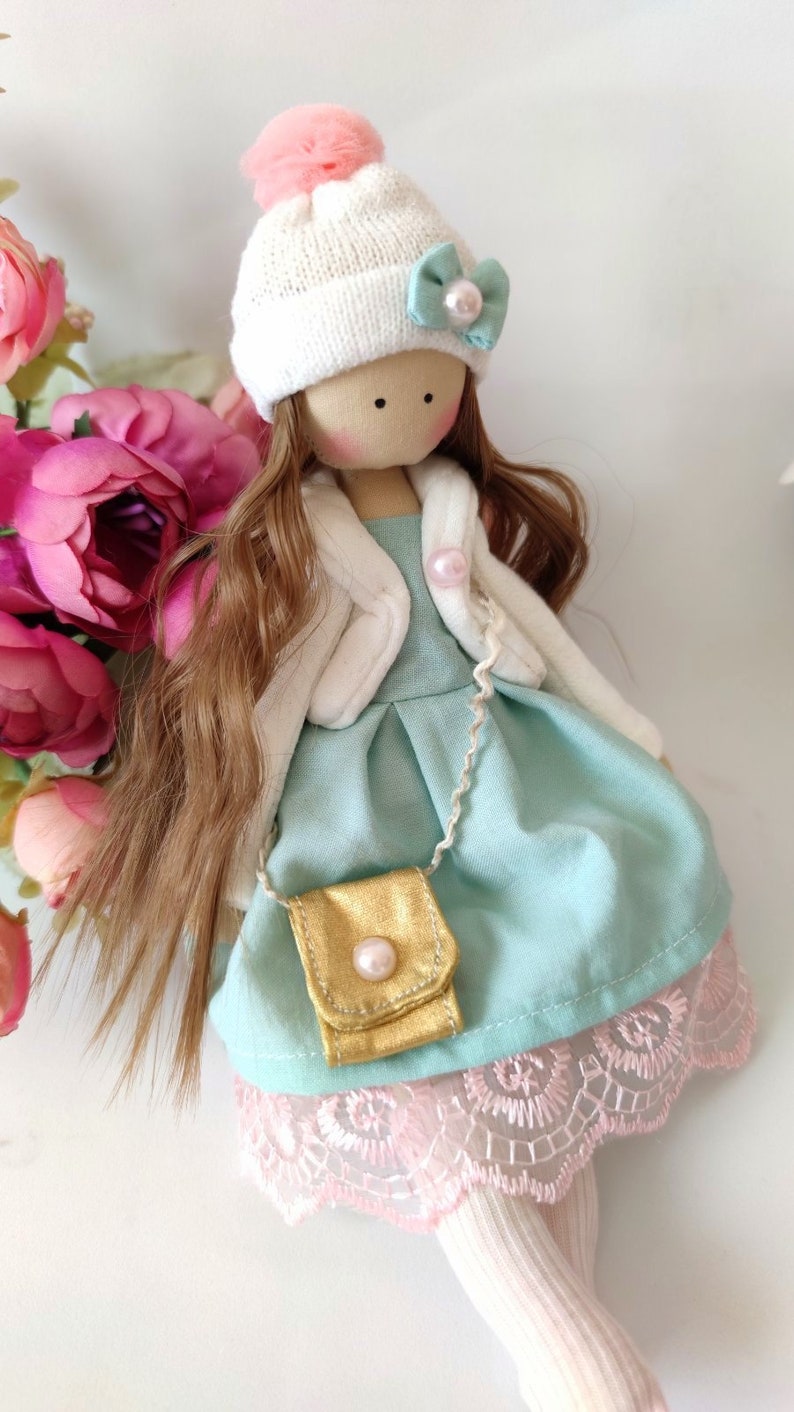 Puede incluir: Una mu&ntilde;eca hecha a mano con cabello largo y casta&ntilde;o, que lleva un gorro blanco de punto con un pomp&oacute;n rosa, una chaqueta blanca, un vestido azul claro con un bolso dorado y calcetines blancos.