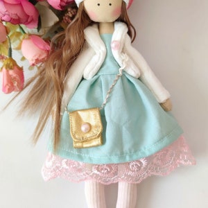 Puede incluir: Una mu&ntilde;eca hecha a mano con cabello largo y casta&ntilde;o, que lleva un sombrero blanco con un pomp&oacute;n rosa, una chaqueta blanca, un vestido azul claro con un bolso dorado y medias rosas.