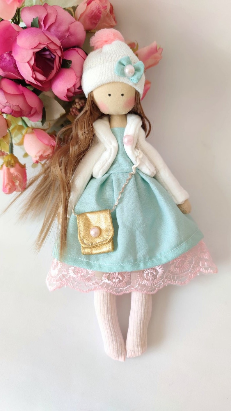 Puede incluir: Una mu&ntilde;eca hecha a mano con cabello largo y casta&ntilde;o, que lleva un sombrero blanco con un pomp&oacute;n rosa, una chaqueta blanca, un vestido azul claro con un bolso dorado y medias rosas.