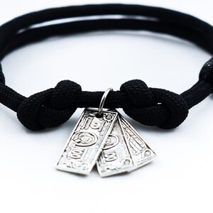 May include: A black paracord bracelet with a silver charm of three stacked dollar bills.