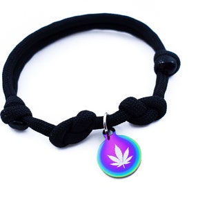 May include: Black paracord bracelet with a silver charm featuring a white marijuana leaf design. The charm has a rainbow iridescent finish.