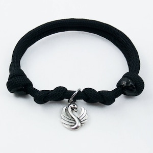 May include: Black rope bracelet featuring a silver swan charm. The bracelet is made from thick, woven black rope, with knots at each end. The swan charm is detailed and hangs from a small silver ring.