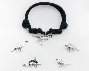 May include: A black paracord bracelet with silver dinosaur charms. The charms include a Tyrannosaurus Rex, a Triceratops, a Stegosaurus, and two other dinosaurs.