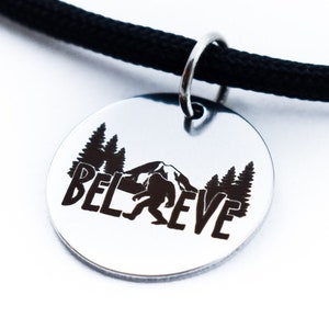 May include: A silver necklace with a black cord and a round pendant. The pendant has a silhouette of Bigfoot walking in front of a mountain and trees, with the word "BELIEVE" engraved below.
