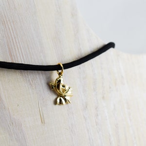 Puede incluir: Un collar con un colgante de p&aacute;jaro dorado en un cord&oacute;n el&aacute;stico negro. El collar se muestra sobre una superficie de madera.
